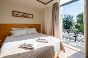 Una camera da letto con testiera in legno, un letto con lenzuola bianche, vista su un balcone con mobili da esterno e un quadro appeso al muro.