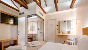 Habitación de hotel con cama, escritorio, silla, espejo y zona de ducha.