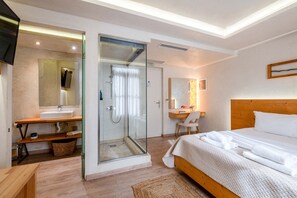 Ett modernt hotellrum med en dusch som är omgiven av glasväggar, en säng och ett skrivbord i trä samt en stor spegel.