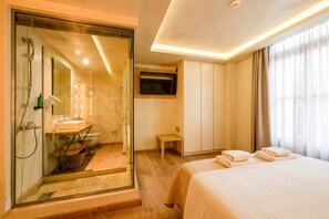A hotel room with a glass shower enclosure, a bed with neatly folded towels, a television mounted on the wall, and a wooden bench. - Panellinion Beachfront Gem Balcony With Cityview (Kalamata)