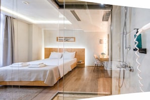 A modern hotel room with a glass partition, a wooden bed, a bedside table, a chair, and a shower area. - Panellinion Beachfront Gem Balcony With Cityview (Kalamata)