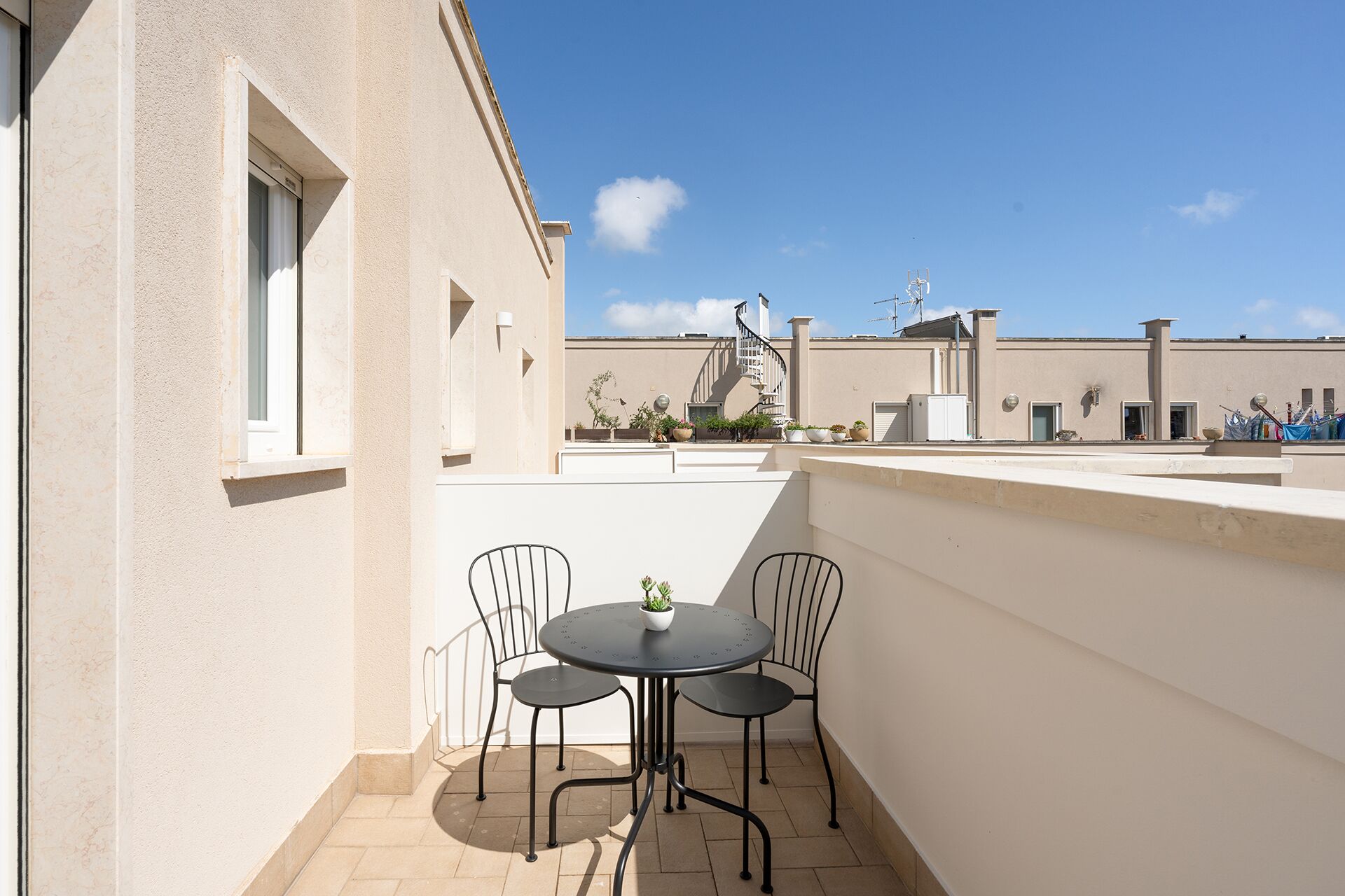 A small outdoor patio with a round table and four chairs, a potted plant, and a view of a building with a spiral staircase.
