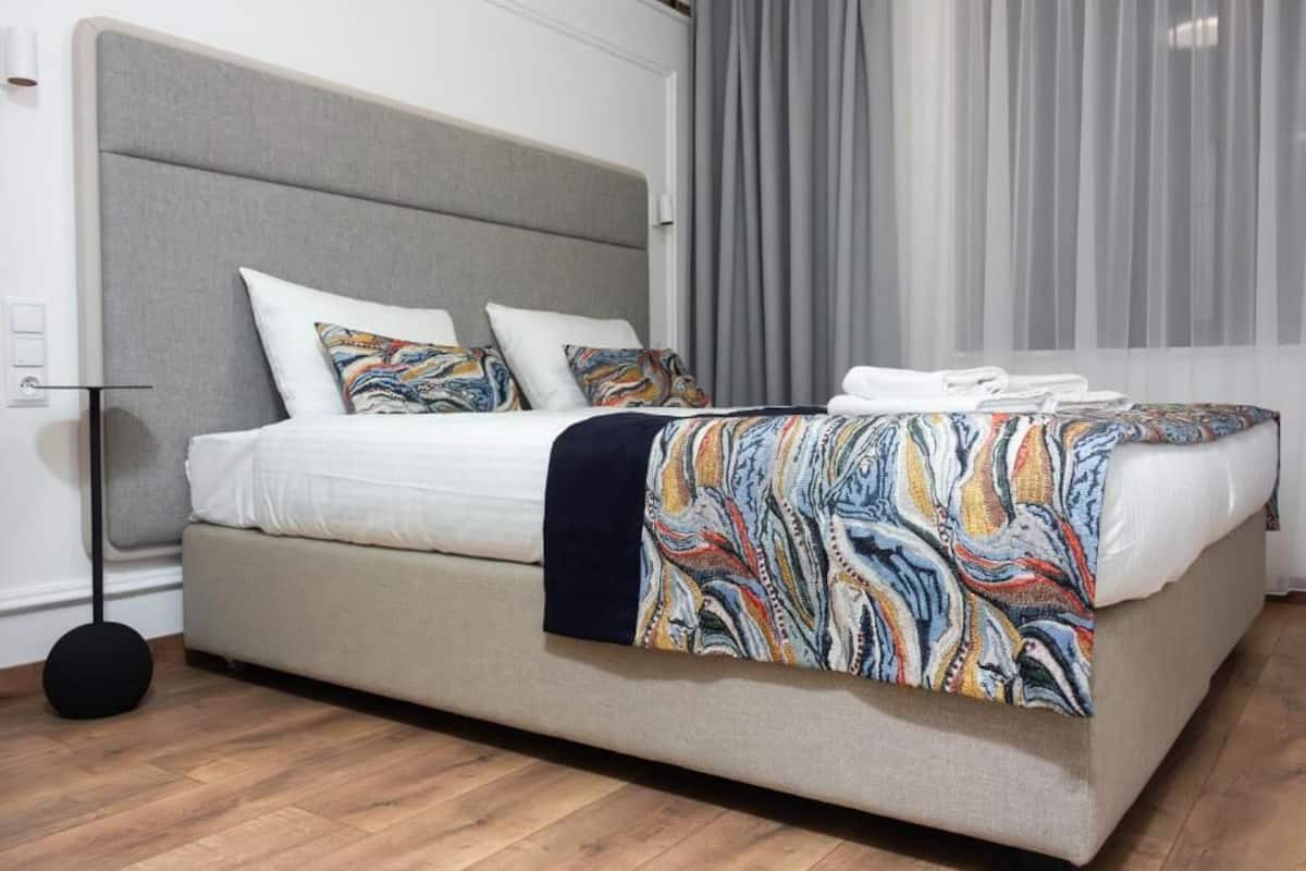 Ein Bett mit grauem Kopfteil, weißer Bettwäsche und einer bunten Decke.
