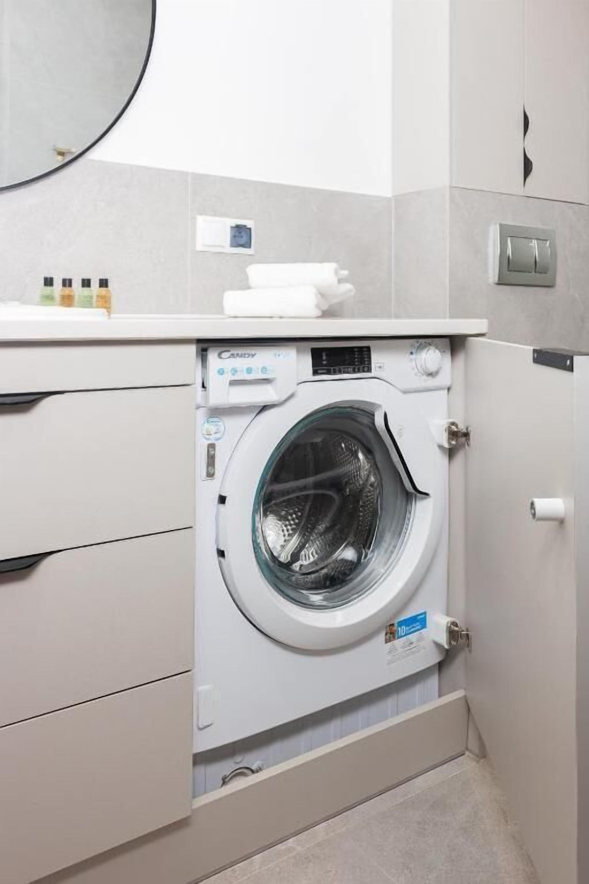 Uma área de lavanderia compacta com máquina de lavar de abertura frontal, um pequeno armário com produtos de higiene e um interruptor de luz na parede.