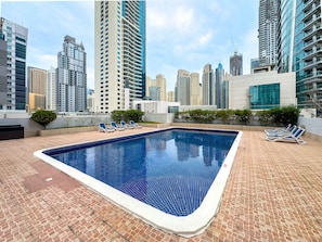 A rooftop pool with blue tiles, surrounded by lounge chairs and a cityscape view. - Free Housekeeping Visits During Your Stay - Stayshort - Chic 1BR in Dubai Marina Sleeps 2 (Dubai)