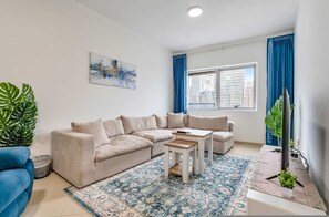 A living room with a beige sectional sofa, a small wooden coffee table, a patterned rug, a large window with blue curtains, and a painting on the wall. - Free Housekeeping Visits During Your Stay - Stayshort - Chic 1BR in Dubai Marina Sleeps 2 (Dubai)