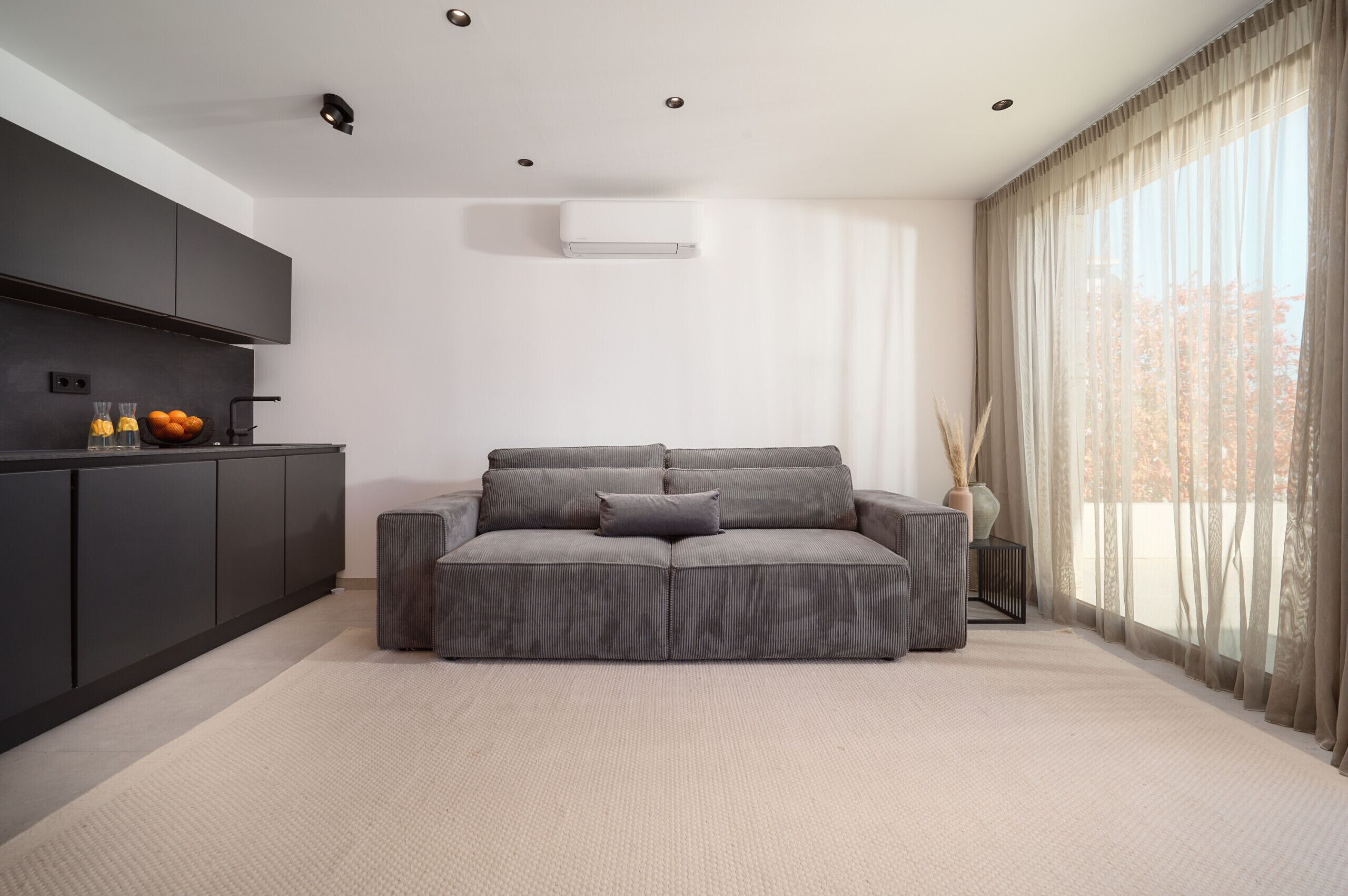 A modern living room with a sofa, a kitchen area, and a large window with curtains.