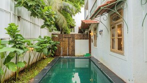 Una piscina con cerda de madera, abundante vegetación y un edificio con tejado de tejas y una ventana con lámpara.