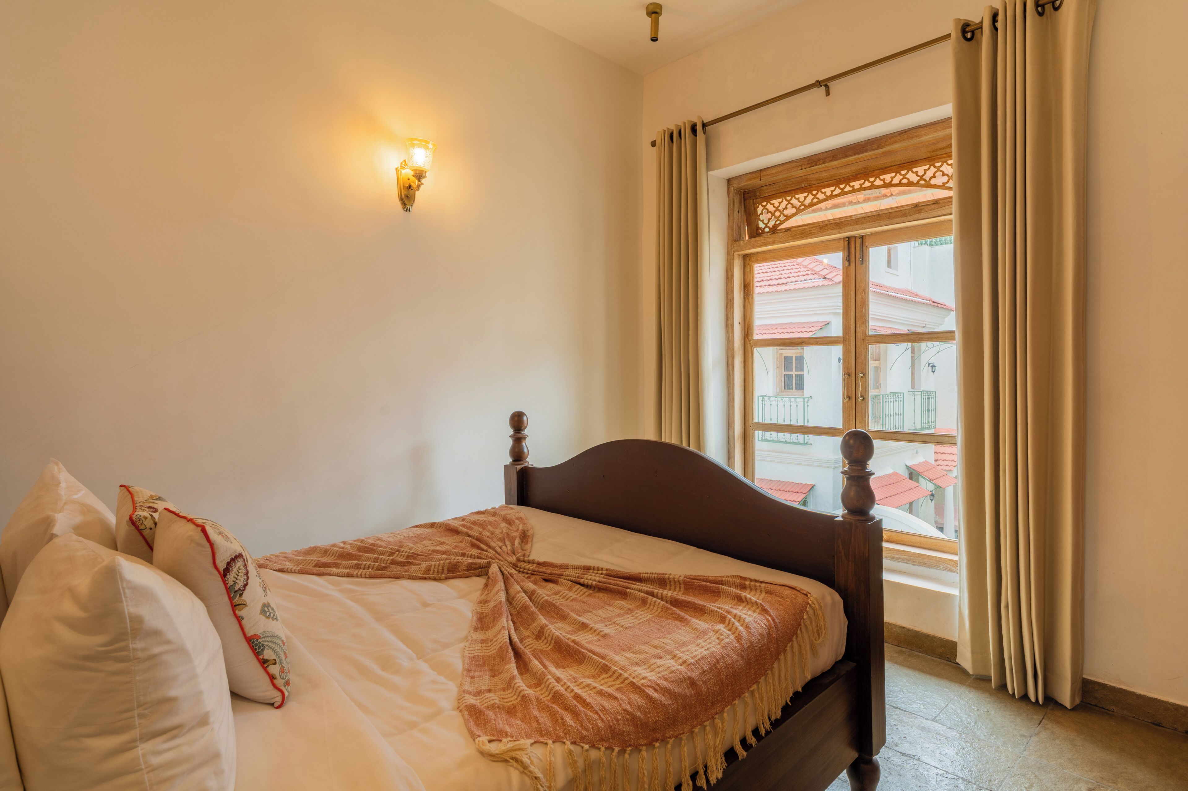 A bedroom with a bed, a wall-mounted light, a window with curtains, and a view of buildings outside.
