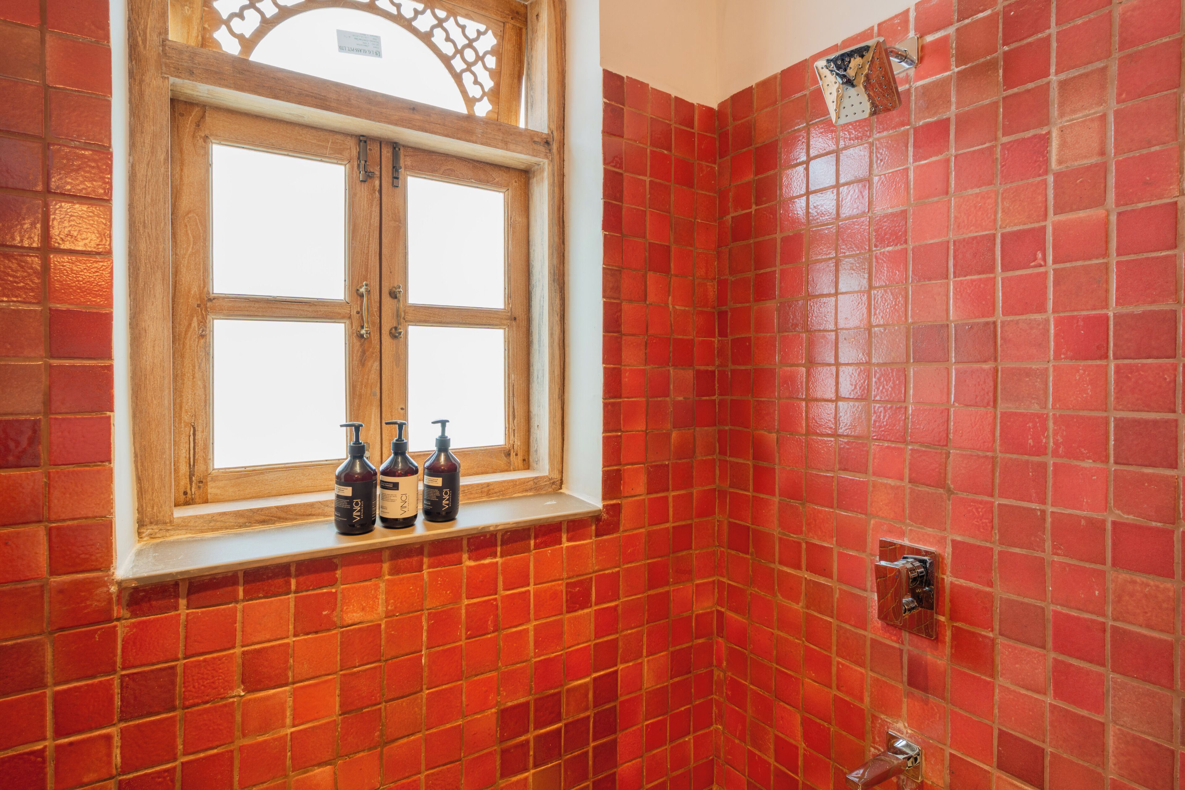 A bathroom with red tiled walls, a wooden window with bottles on the sill, and a shower area.