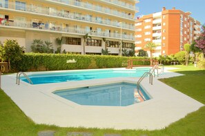 Una piscina amb una zona enjardinada al voltant, un edifici amb balcons i una tanca ben cuidada.