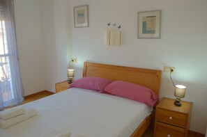 A bedroom with a wooden bed, two bedside lamps, a nightstand with a lamp, a window with sheer curtains, and wall-mounted light switches. - Jasmin Apartment for 4 guests, air conditioning   (Lloret de Mar)