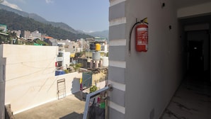 A building with a fire extinguisher mounted on the wall, a visible door, and a view of a residential area with houses and mountains in the background. - Hotel Parth Inn (Rishikesh)