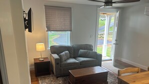 Una sala de estar con un sofá gris, una mesa de centro de madera, una lámpara y vistas al exterior a través de una ventana.
