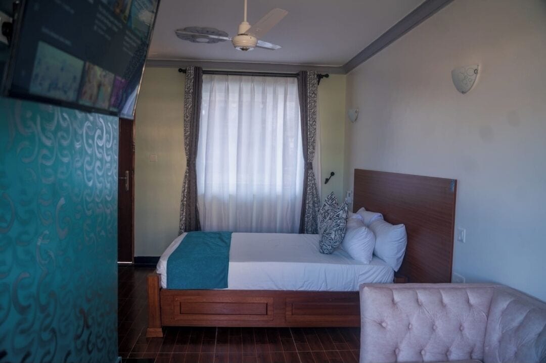 A bedroom with a bed, a ceiling fan, curtains, a wall-mounted light, and a patterned wall.