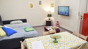 Un dormitorio con cama, una mesa con mantel floral, un televisor en la pared y una puerta.