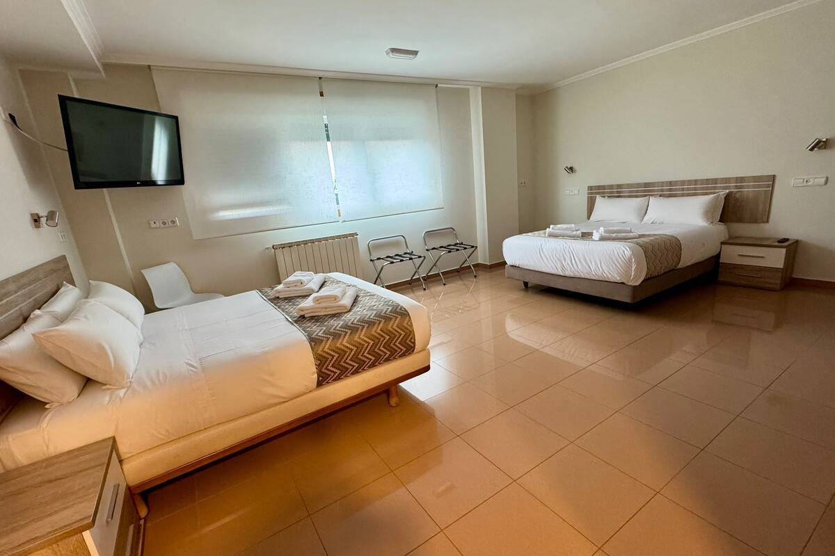 A hotel room with two beds, a TV mounted on the wall, a window with blinds, and a radiator.