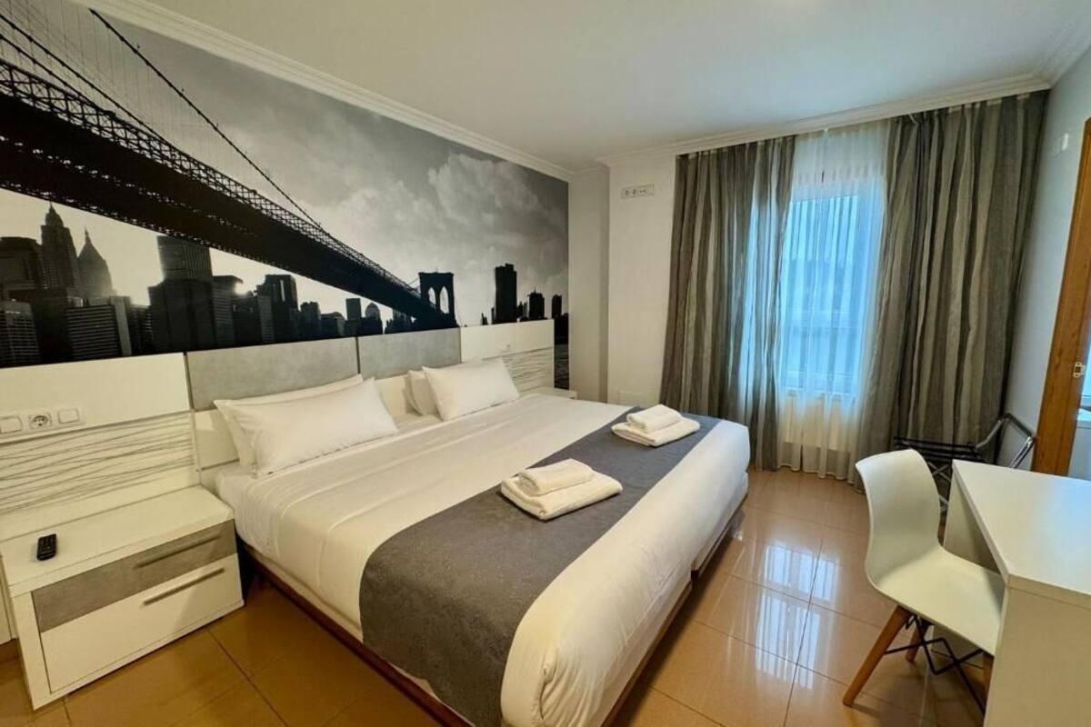 A hotel room with a bed, a desk, a chair, a nightstand, and a cityscape mural on the wall.