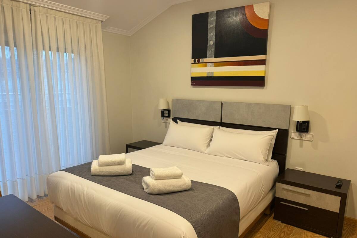 A hotel room with a bed, two bedside tables, a nightstand, a desk, and a painting on the wall.