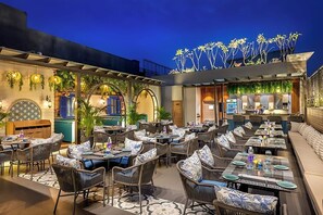 Un área de comedor al aire libre con sillas y mesas de mimbre, una zona de bar con mostrador y un techo decorativo con motivos vegetales.