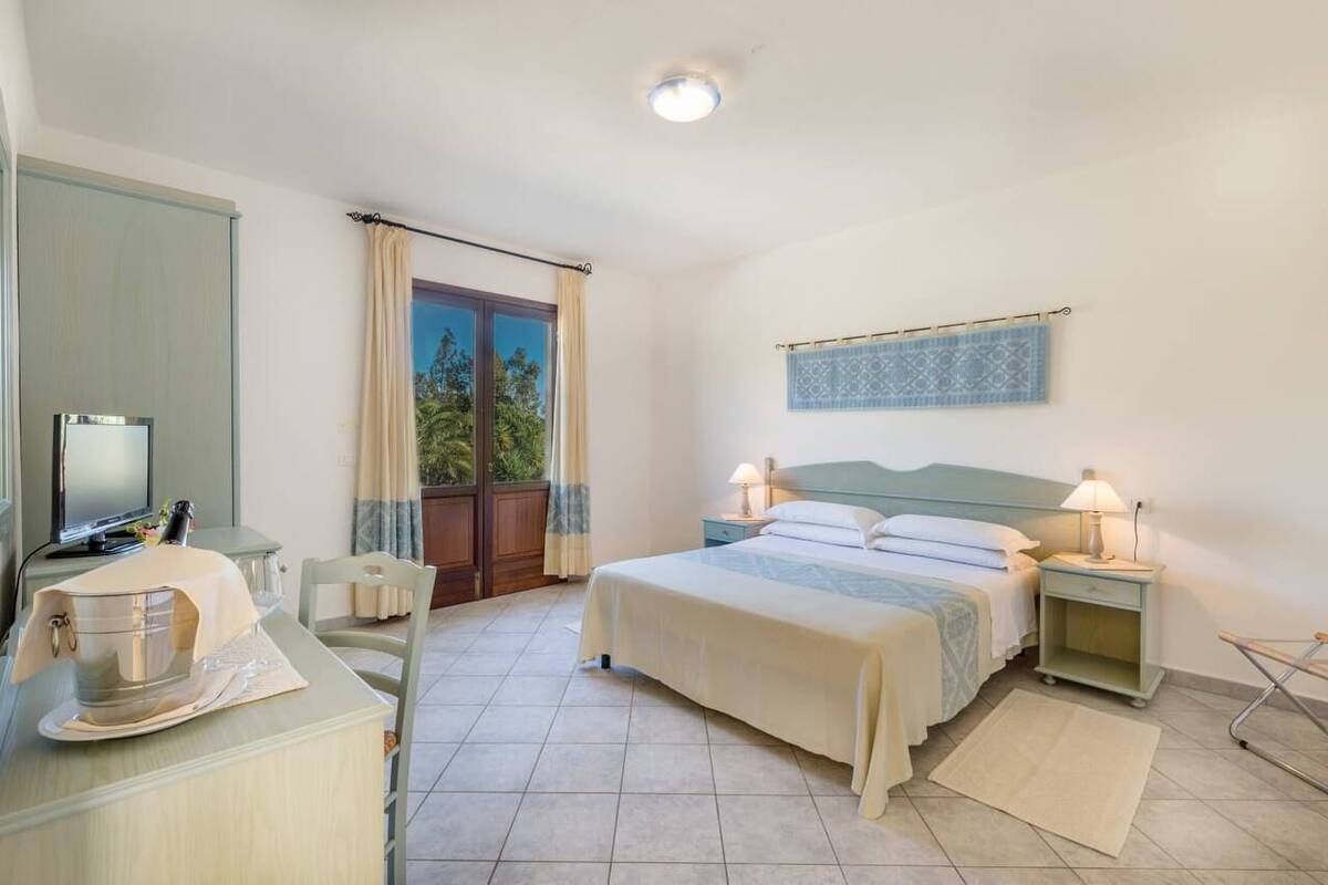 Une chambre à coucher avec un lit, des tables de chevet, un bureau, une télévision et une fenêtre avec des rideaux.