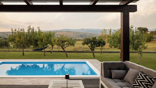 Un espace piscine agrémenté d’un canapé et d’une lampe, entouré d’une terrasse en bois et offrant une vue sur un paysage vallonné.