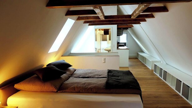 Ein Schlafzimmer mit schrägen Wänden, einem Bett mit Kissen, einem Nachttisch und einem Fenster.
