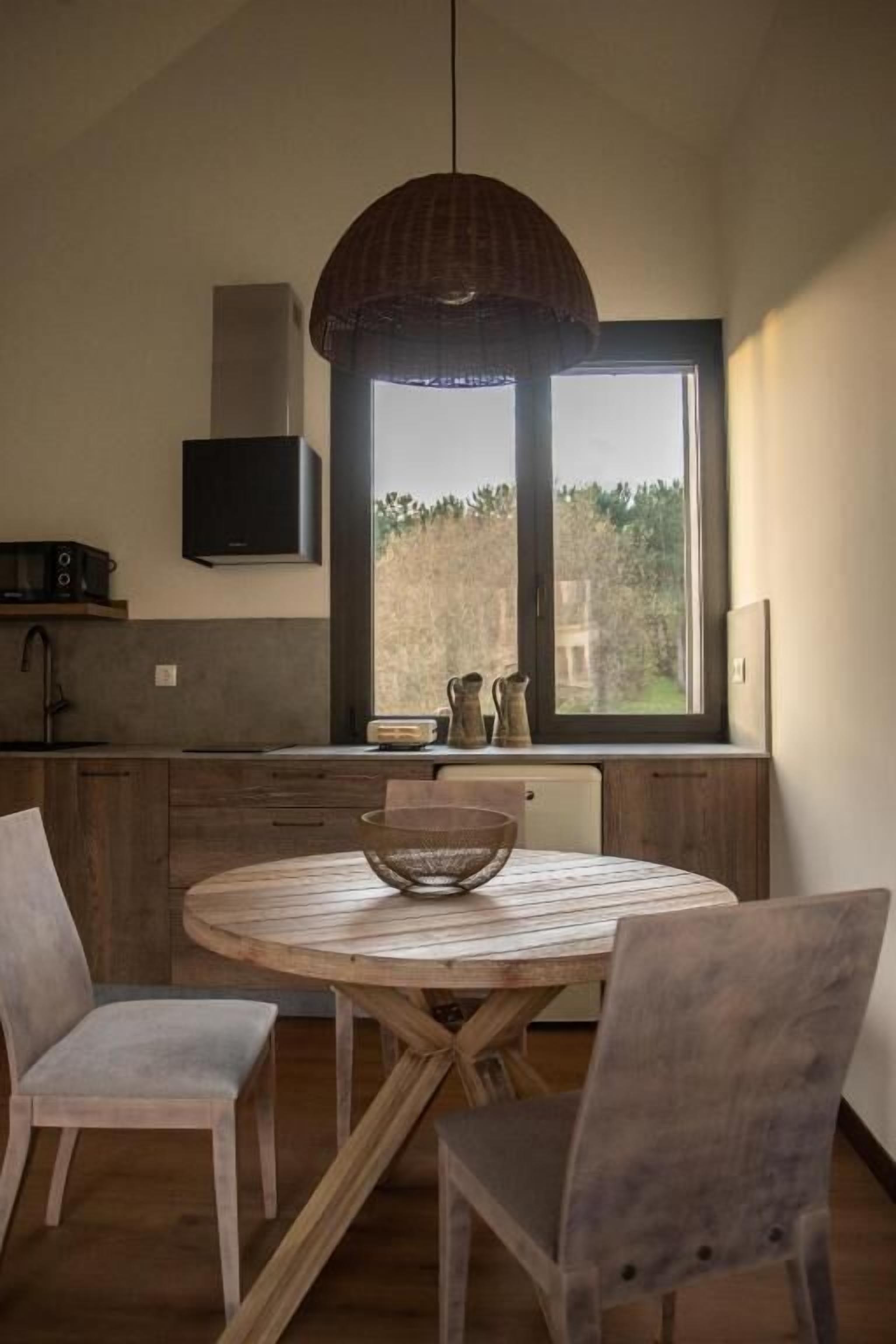 A modern kitchen with a round wooden table, two chairs, a hanging light fixture, and a window with a view of greenery.