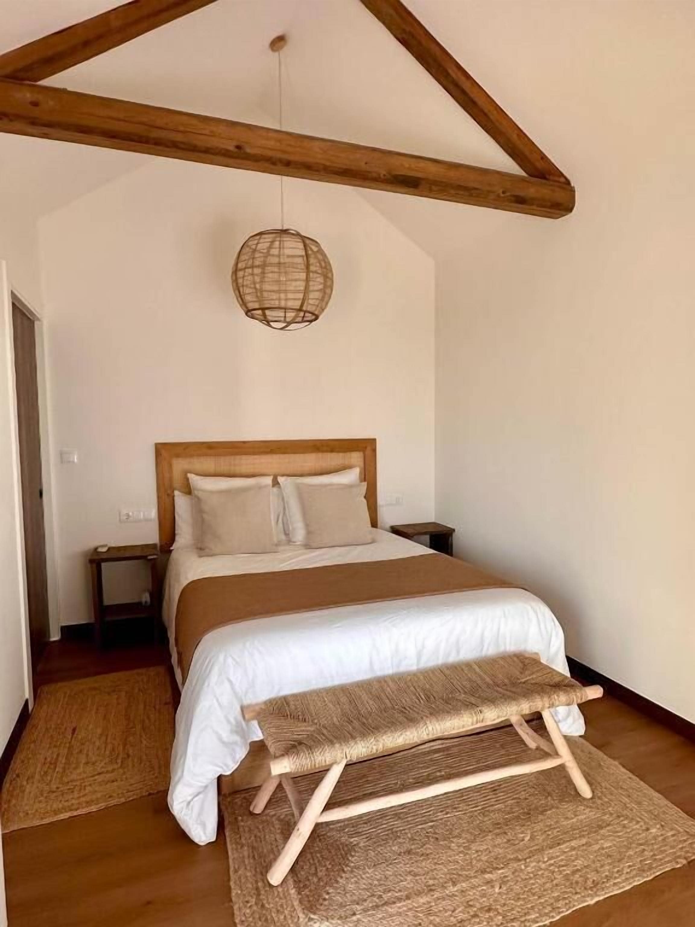 Un dormitorio con techo de madera, una cama con cabecera, una mesita de noche, una lámpara colgante y un banco.