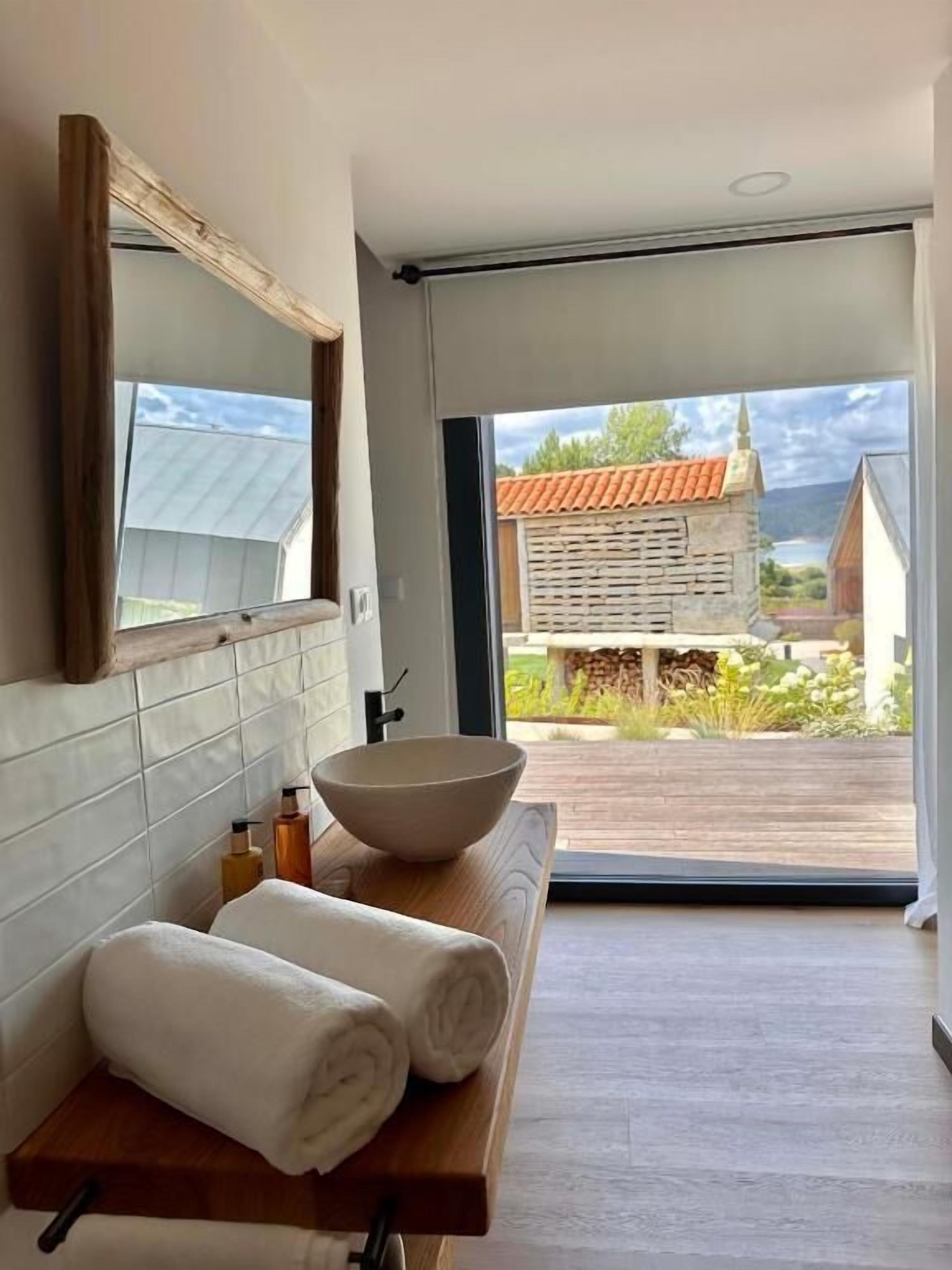 A bathroom with a large mirror, a wooden vanity, a bowl sink, and a view of a house through a glass door.