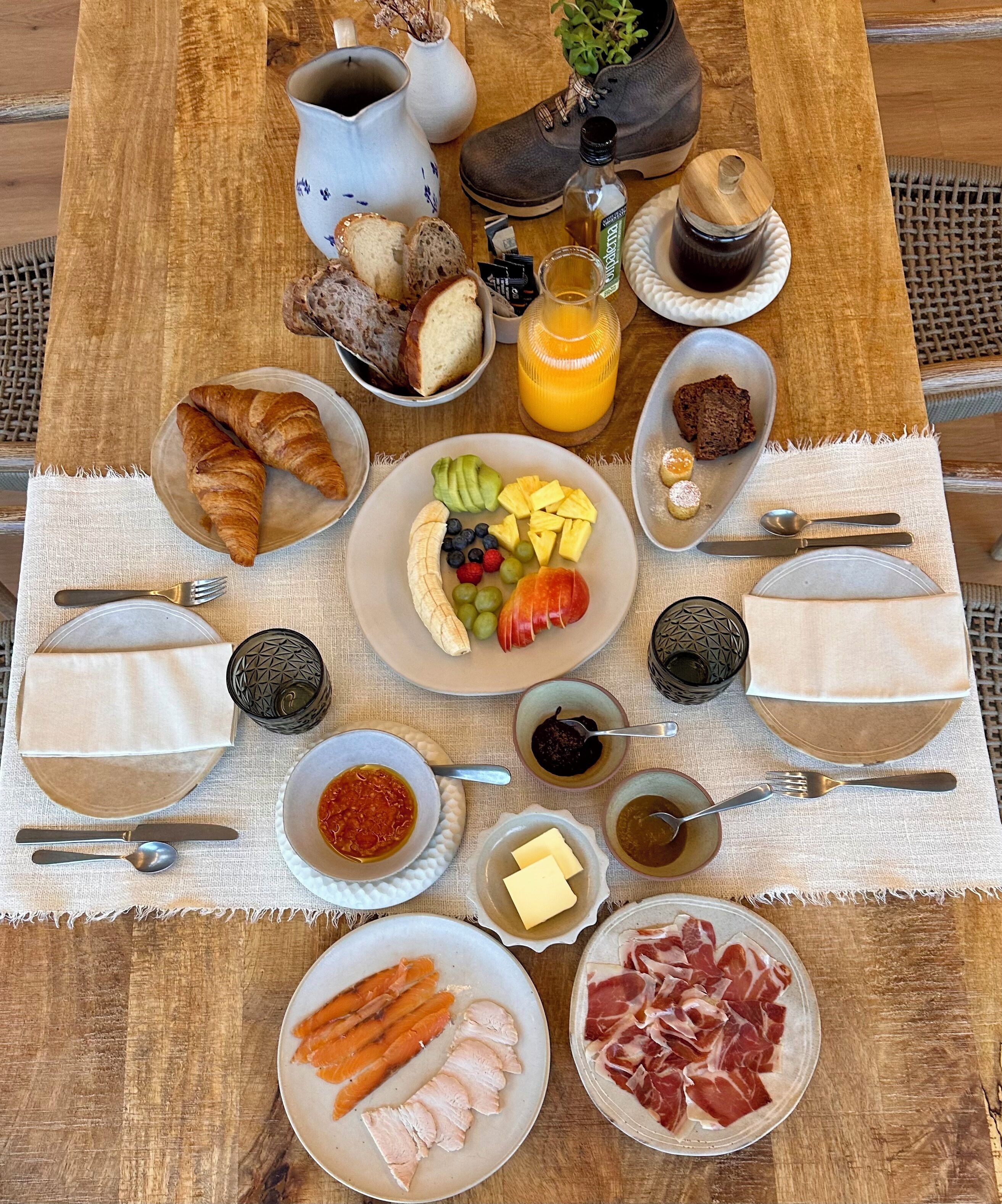 Une table en bois sur laquelle sont disposés divers aliments pour le petit-déjeuner, notamment des croissants, des fruits et de la charcuterie, le tout présenté sur un set de table en fibres tressées.