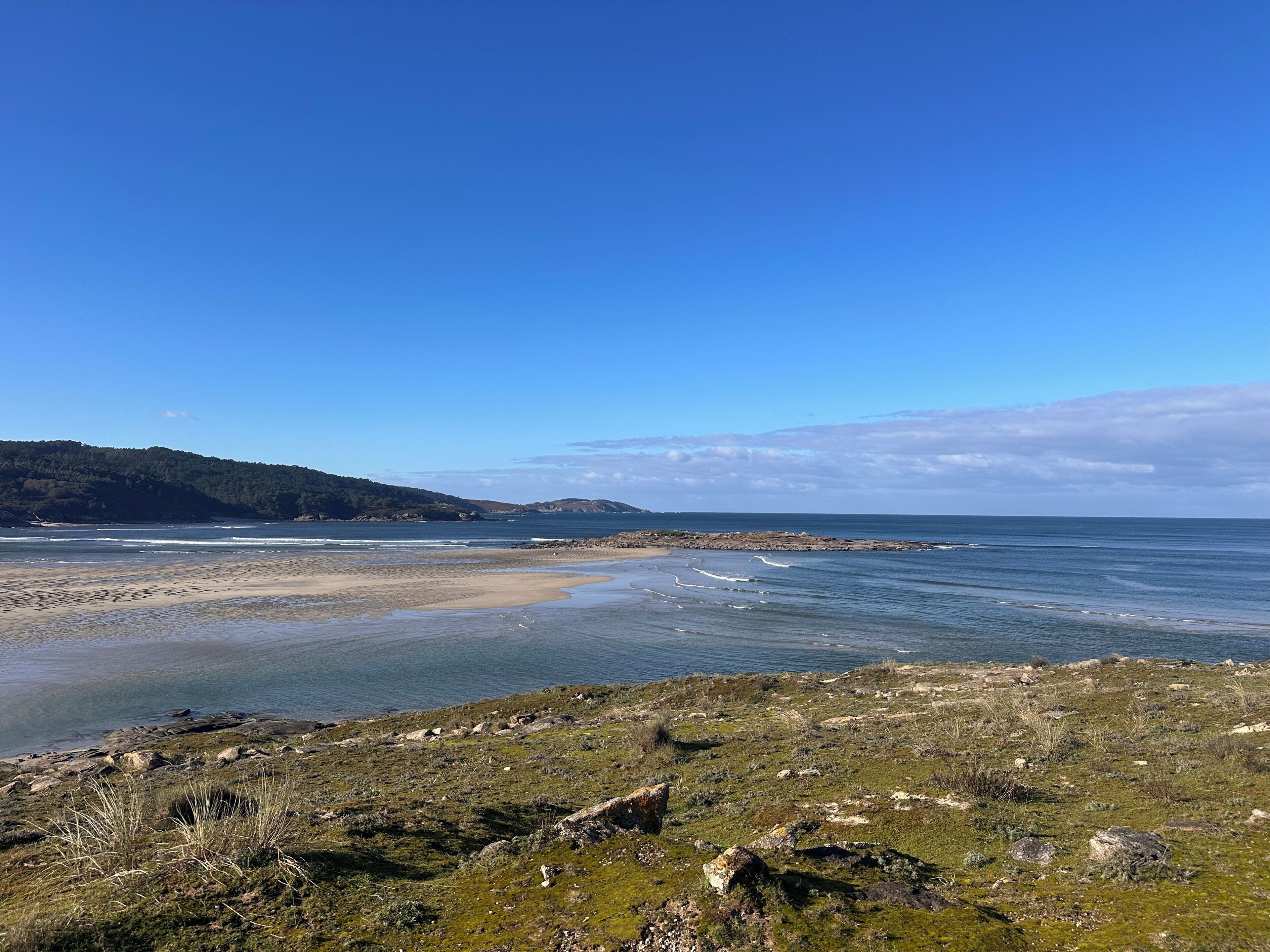A coastal landscape with a beach, rocky shoreline, and distant hills.