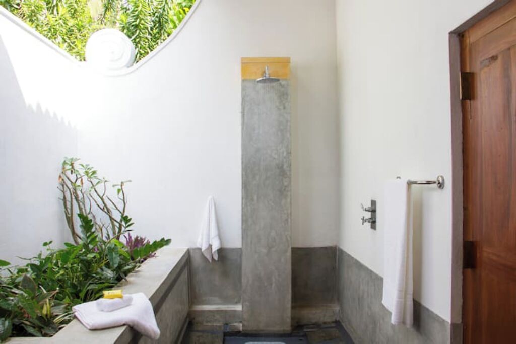 Un petit espace de douche fermé, avec une colonne en béton, une porte en bois et une fenêtre donnant sur un espace verdoyant.