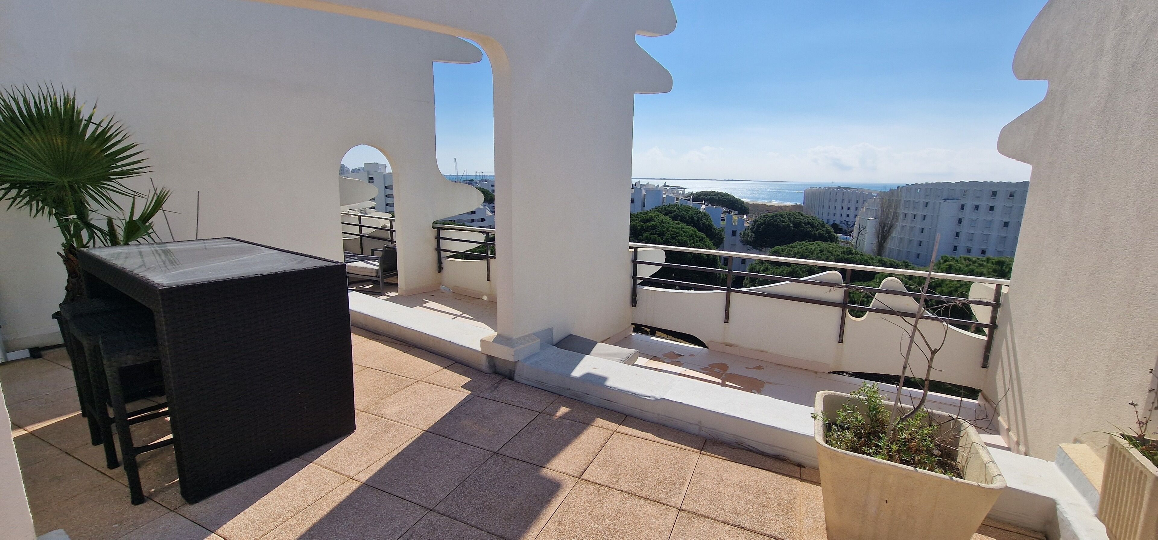 A balcony with a table and chairs, potted plants, and a view of buildings and the sea.
