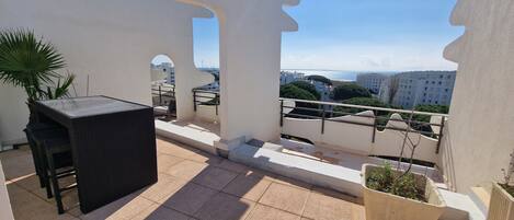 A balcony with a table and chairs, potted plants, and a view of buildings and the sea.