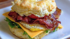 A breakfast sandwich with a biscuit, bacon, egg, cheese, and tomato on a white plate. - Hotel Air Admire - IGI Airport Delhi (New Delhi)