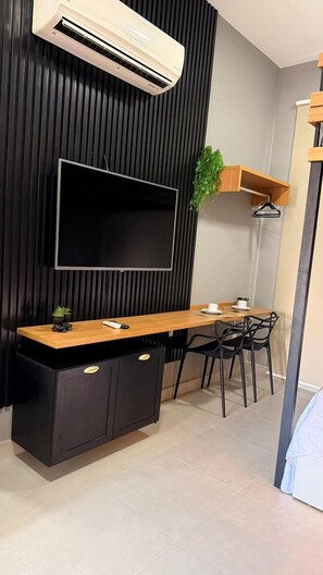 A modern room with a flat-screen TV mounted on a wall, a wooden desk with a black cabinet, a dining area with chairs, and a small plant on the wall. - Beach Flat Sonne (João Pessoa)