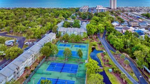 Luchtbeeld van een woongebied met meerdere tennisbanen, een groot gebouw met een plat dak en een waterpartij op de achtergrond.
