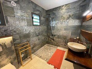 A bathroom with stone walls, a shower, a wooden-framed shelf, a red rug, and a large sink.