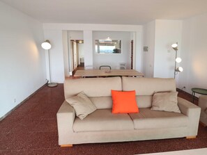 Ein sofas in neutralen Farben mit einem einzelnen orangefarbenen Kissen, ein Esstisch mit Stühlen und zwei Stehlampen in einem modernen, gut beleuchteten Raum.