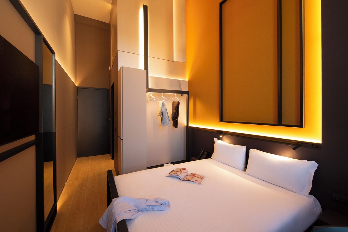A hotel room with a bed, a wardrobe, a mirror, and a wall-mounted light fixture.