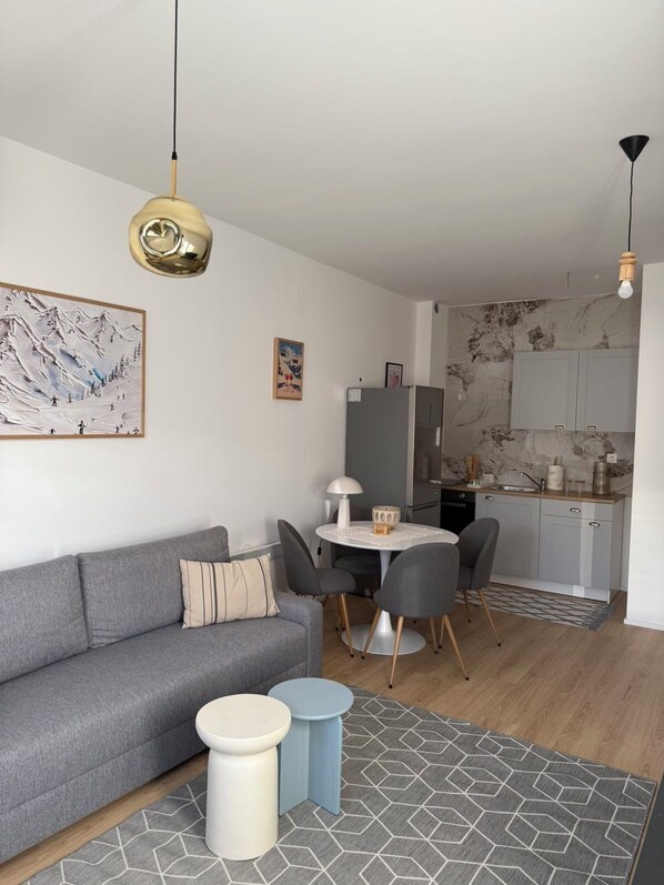 A modern living room with a grey sofa, a dining table with chairs, a kitchen area with marble backsplash, and a patterned rug.