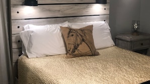 A bed with white linens, a textured beige blanket, and a horse-themed pillow. There are two black wall-mounted lamps on the headboard. - Louisville's Central "Lilly Pad" (Louisville)