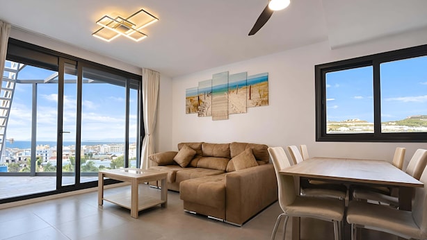 A modern living room with a sofa, coffee table, dining table, and chairs. There are large windows with a view of the city. - Marinsa Beach - Penthouse (Torrox)