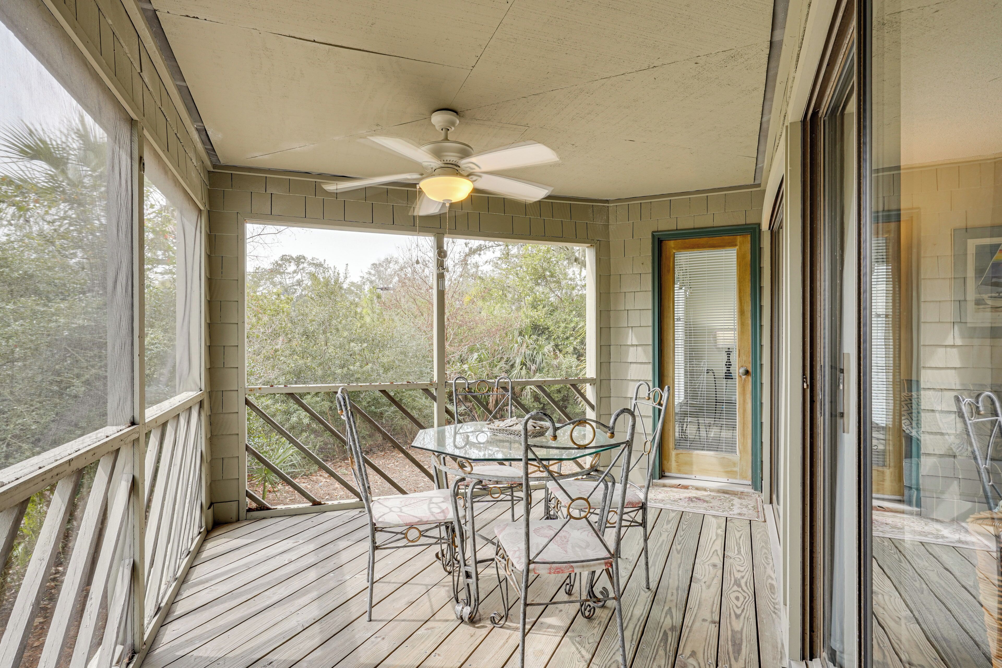 Une terrasse extérieure couverte, avec une table et des chaises, un ventilateur de plafond et une vue sur les arbres à travers les fenêtres.