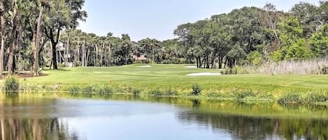 En golfbana med en damm i förgrunden, omgiven av träd och en klar himmel.