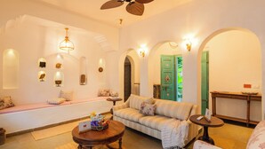 A living room with a sofa, a round coffee table, and a bench. There are wall sconces, a ceiling fan, and a green door. - ELIVAAS Amayah Rasa 4 BHK Villa (Assagao)
