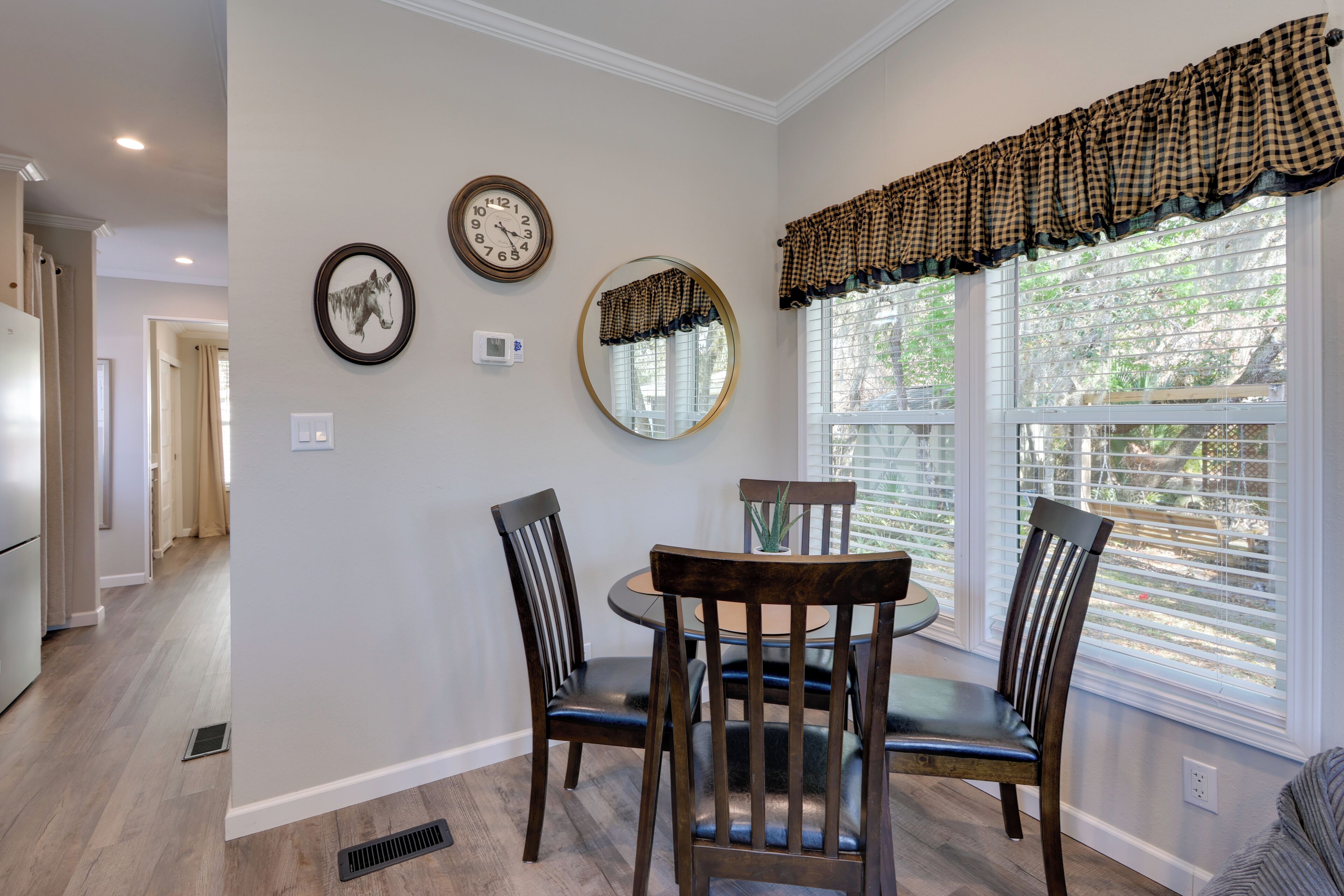 A dining area with a round table, four chairs, a wall clock, a mirror, and a window with blinds.
