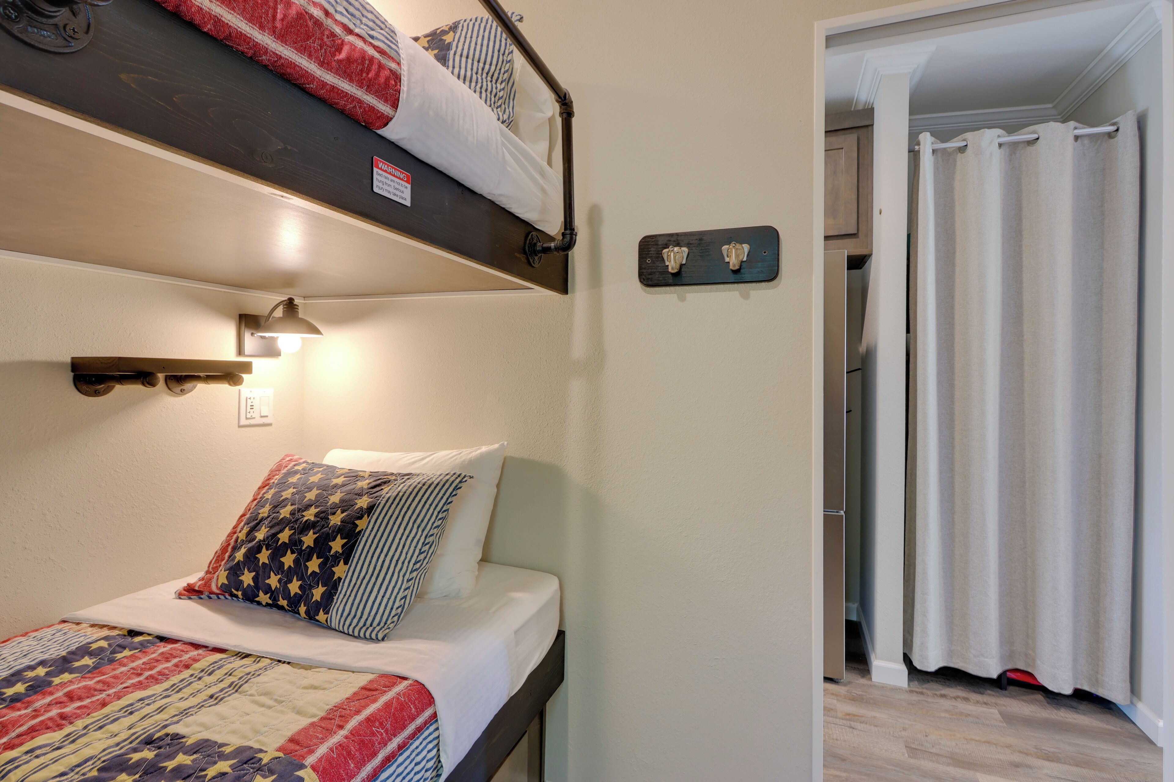 Ein Etagenbett mit Kopfteil, eine Nachttischlampe und eine Steppdecke. Eine Tür mit Vorhang und ein an der Wand befestigter Lichtschalter.