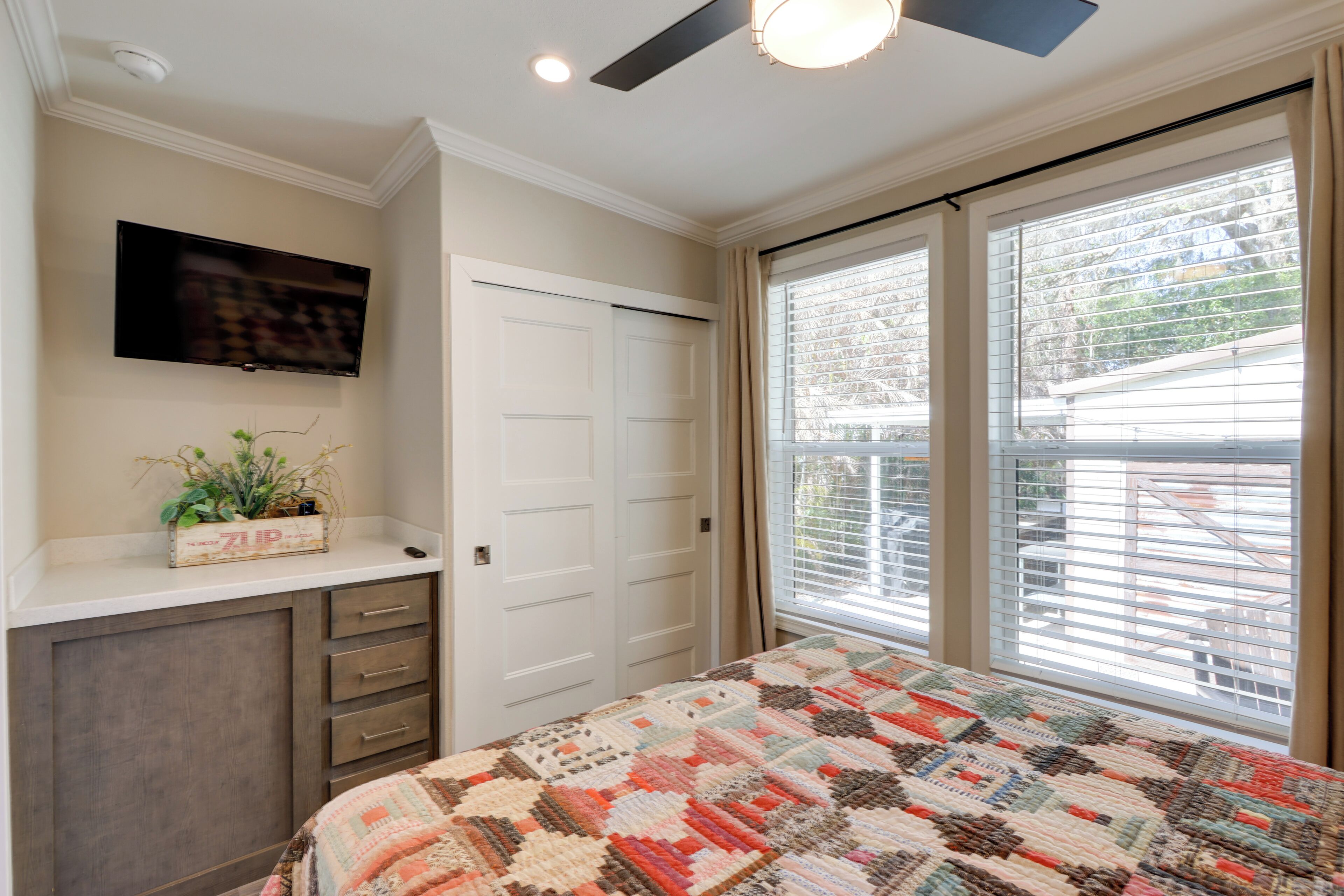 A bedroom with a bed, a TV, a ceiling fan, a window with blinds, and a dresser.